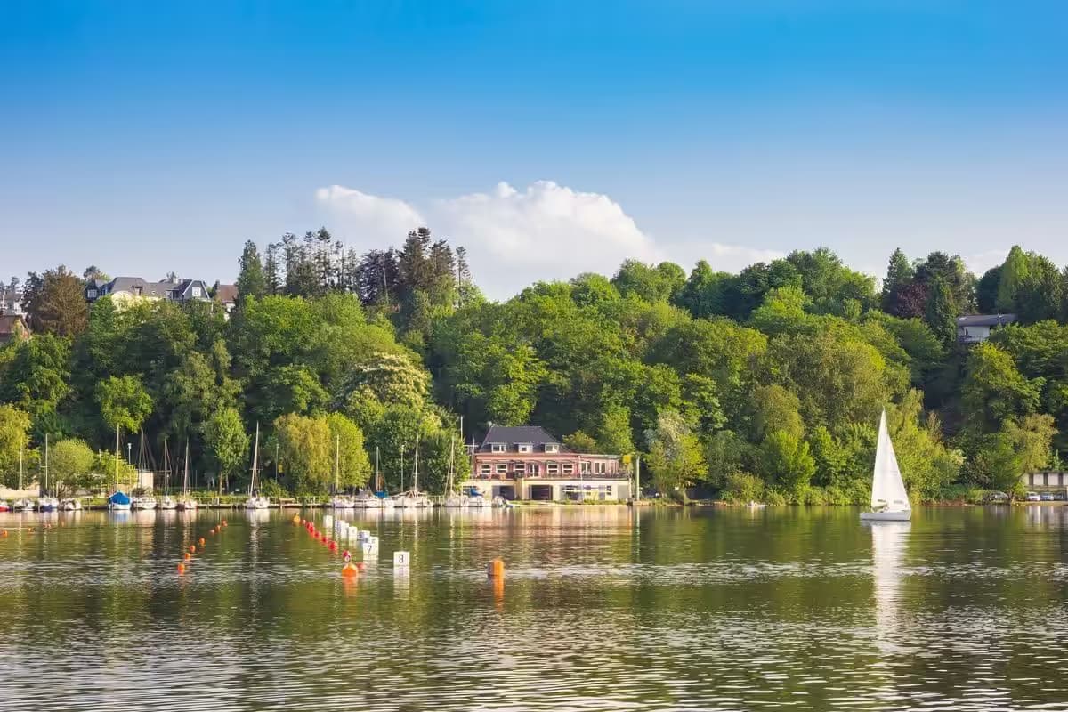 Segelboot auf ruhigem See vor bewaldeter Uferlandschaft.