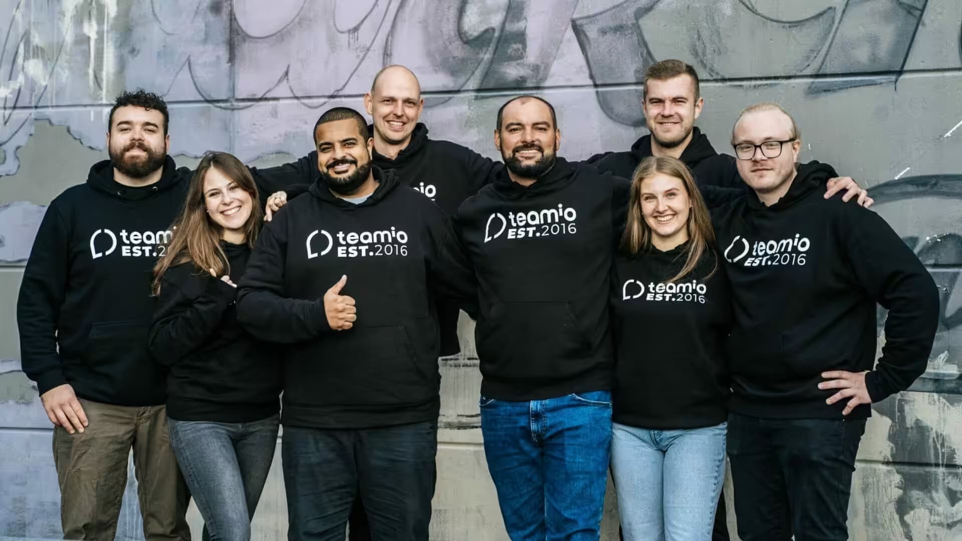 Das teamio-Team in schwarzen Team-Hoodies vor einer Graffitiwand