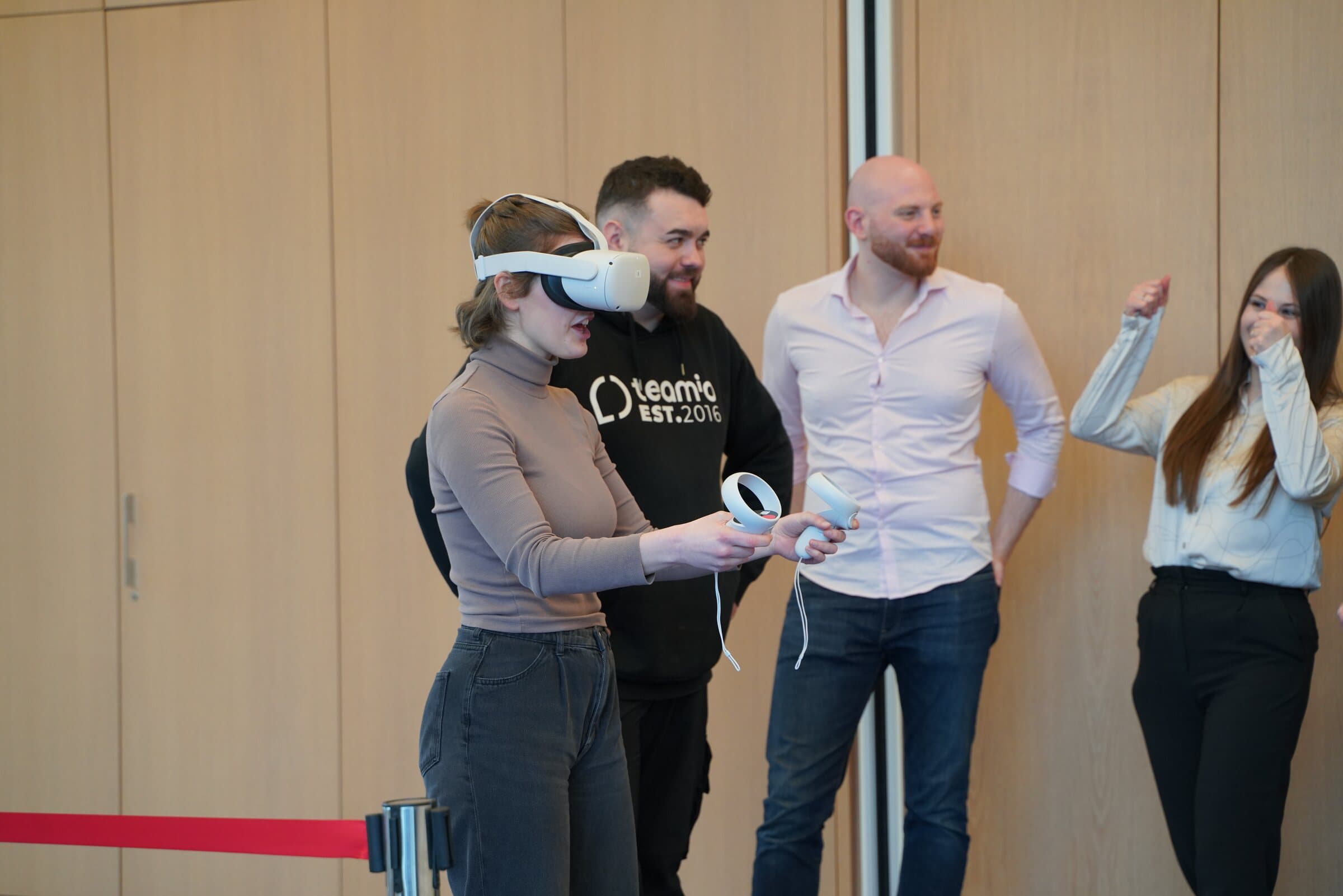 Teilnehmerin mit VR-Brille und Controllern, Kollegen schauen lachend zu
